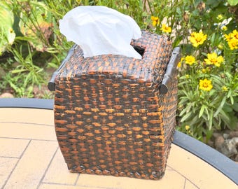 May include: A square tissue box cover crafted from woven dark brown and black material with a glossy finish. The box features a rectangular opening at the top with a white tissue protruding from it and dark, bamboo-style accents on the sides.