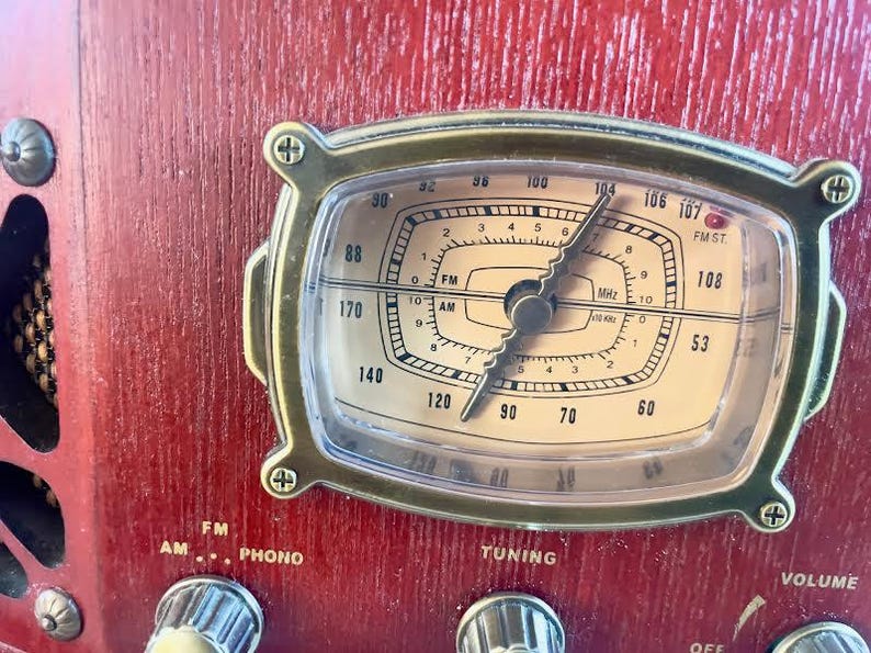 May include: Close-up of a vintage-style radio with a red wooden cabinet. The radio dial is framed in gold and features a clear display with numbers and a needle. Knobs for tuning and volume are visible.
