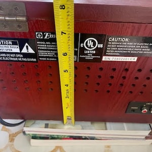 May include: A vintage, dark red wooden radio with a yellow measuring tape showing a height of approximately 18 cm. The radio has several stickers with text, including a UL listing and a warning about electric shock.