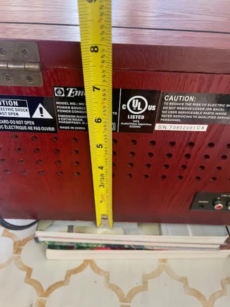 May include: A vintage, dark red wooden radio with a yellow measuring tape showing a height of approximately 18 cm. The radio has several stickers with text, including a UL listing and a warning about electric shock.
