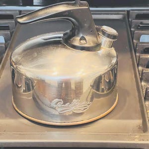 May include: A polished stainless steel Paul Revere tea kettle with a black handle and a matching black whistle cap. The kettle features a cylindrical body with a copper-toned base rim and the Paul Revere signature embossed on the side of the metal.