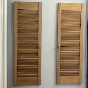 Op de afbeelding: Twee houten luiken met horizontale lamellen zijn aan een lichtgrijze muur bevestigd. De luiken zijn warmbruin van kleur en hebben kleine, ronde zilveren knoppen. De luiken zijn een decoratief element.
