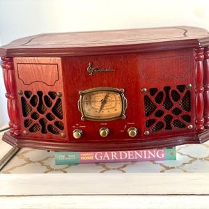 May include: A vintage-style, red wooden radio with a decorative design. The radio features a dial, knobs, and speaker grilles. The radio is sitting on a white table with a book titled "Gardening" underneath.