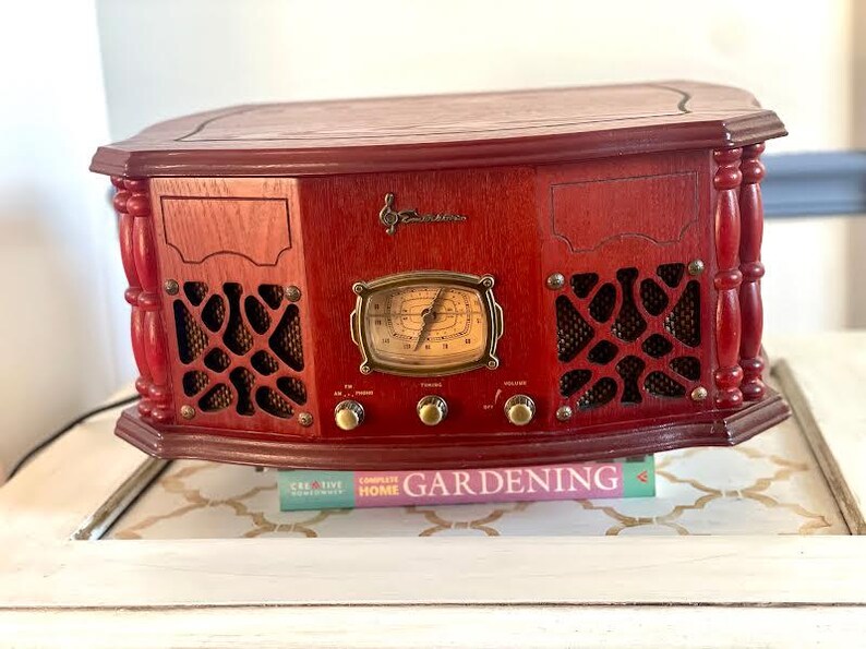 May include: A vintage-style, red wooden radio with a decorative design. The radio features a dial, knobs, and speaker grilles. The radio is sitting on a white table with a book titled "Gardening" underneath.