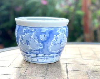 May include: A round ceramic planter featuring a white base with hand-painted blue illustrations of various fruits and foliage. The design includes a repeating pattern of pears and apples around the exterior, finished with a solid white rim.