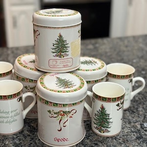 Op de afbeelding: Een verzameling Spode-kerstartikelen, waaronder witte mokken en blikken. De mokken en blikken hebben een kerstboomontwerp, hulst en de tekst "Wishing your Christmas is filled with delights."
