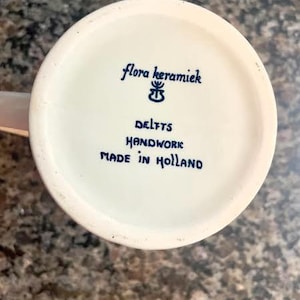 May include: A close-up of the bottom of a white ceramic mug. The text "flora keramiek DELFTS HANDWORK MADE IN HOLLAND" is written in blue script. The mug is on a speckled brown surface.