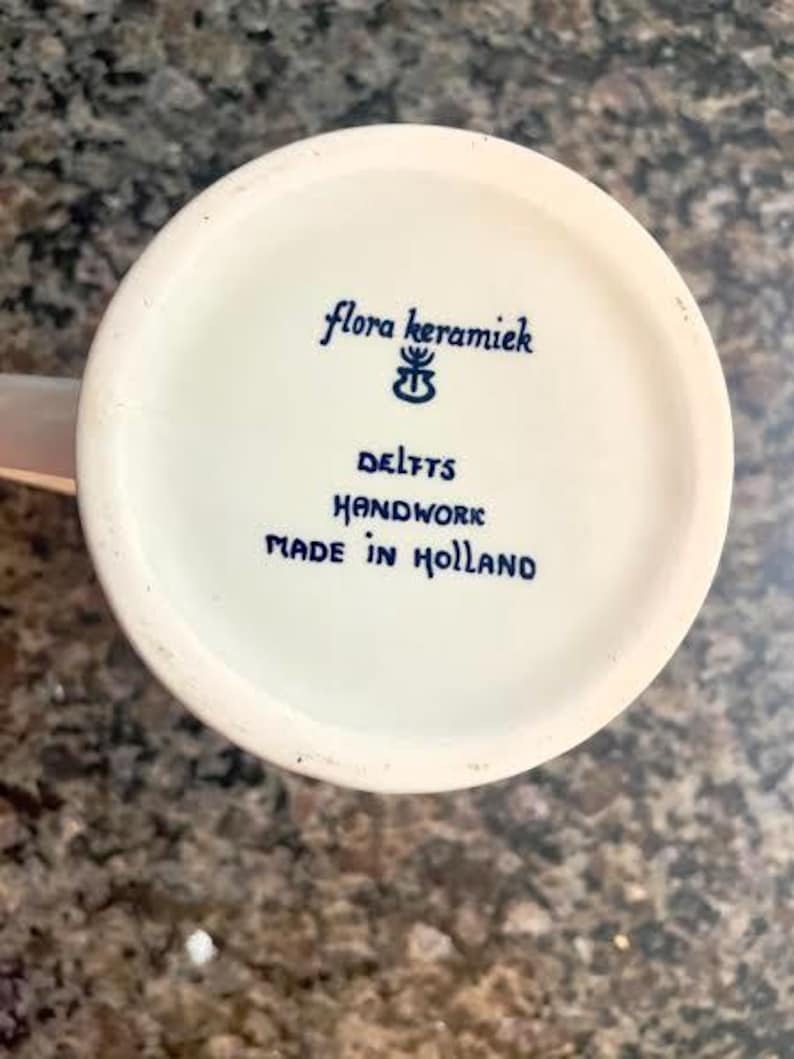 May include: A close-up of the bottom of a white ceramic mug. The text "flora keramiek DELFTS HANDWORK MADE IN HOLLAND" is written in blue script. The mug is on a speckled brown surface.