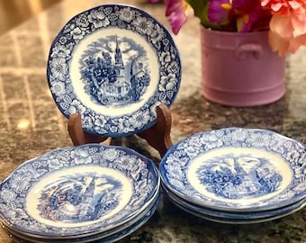 May include: A collection of blue and white ceramic plates. Each plate features a central image of a church and is surrounded by a floral border. The plates are displayed on a table, with one plate on a wooden stand.