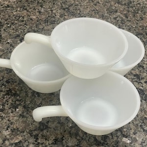 May include: Four white, vintage-style, milk glass bowls with handles. The bowls are stacked and arranged on a speckled, dark gray surface. The bowls have a simple, classic design.