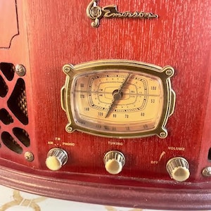 May include: Close-up of a vintage red Emerson radio. The radio features a gold-framed dial, control knobs, and the Emerson logo. The dial displays numbers and a clock-like design. The radio has FM, AM, Phono, Tuning, and Volume controls.