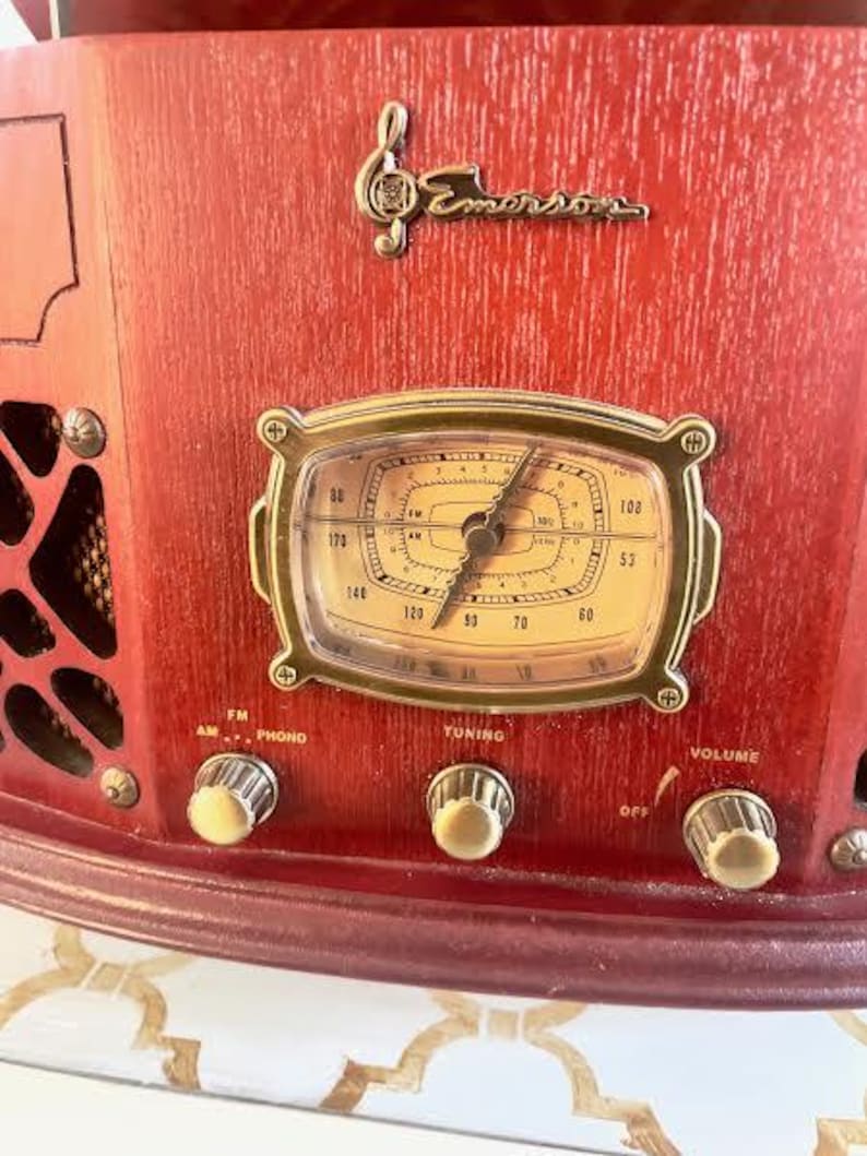 May include: Close-up of a vintage red Emerson radio. The radio features a gold-framed dial, control knobs, and the Emerson logo. The dial displays numbers and a clock-like design. The radio has FM, AM, Phono, Tuning, and Volume controls.