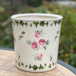 Może przedstawiać: Cylindryczna ceramiczna doniczka o kremowej podstawie, ozdobiona powtarzającym się wzorem różowych skupisk róż i zielonych pnączy bluszczu. Projekt zawiera ciągłą obwódkę z bluszczu wzdłuż górnej i dolnej krawędzi naczynia.
