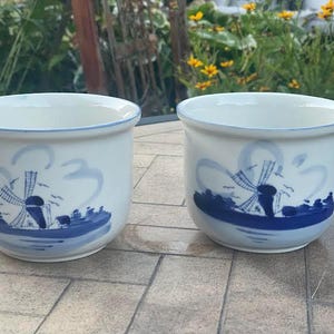 May include: Two white ceramic pots with a blue rim and a blue windmill design. The design features a windmill, water, and clouds. The pots are sitting on a tiled surface.