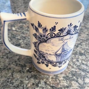 May include: Cream-colored ceramic mug with a blue and white nautical design. The mug features a sailboat scene and floral accents. The handle and rim are also trimmed in blue. The word "Heineken" is visible on the mug.