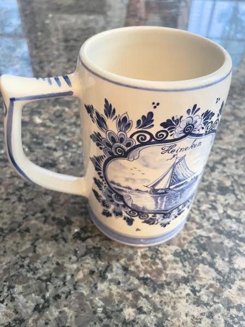 May include: Cream-colored ceramic mug with a blue and white nautical design. The mug features a sailboat scene and floral accents. The handle and rim are also trimmed in blue. The word "Heineken" is visible on the mug.