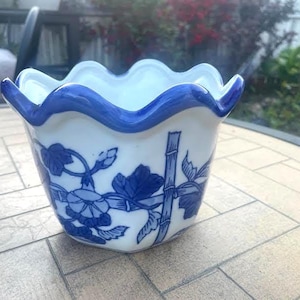 May include: A white ceramic planter with a scalloped rim, accented with a vibrant blue trim. The planter features a detailed blue floral and bamboo design. Ideal for small plants or decorative use.