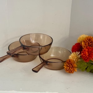 Puede incluir: Dos sartenes redondas de vidrio marrón translúcido con mangos integrados. Las sartenes presentan un acabado liso y pulido con un diseño de cuenco circular poco profundo. Estos recipientes de vidrio están colocados juntos, mostrando su color ámbar uniforme.
