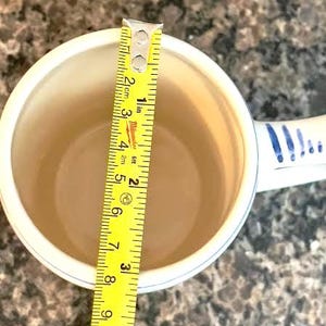 May include: A white ceramic mug with a blue patterned handle and rim, measured with a yellow tape measure. The tape measure shows the mug is approximately 3 inches in diameter.
