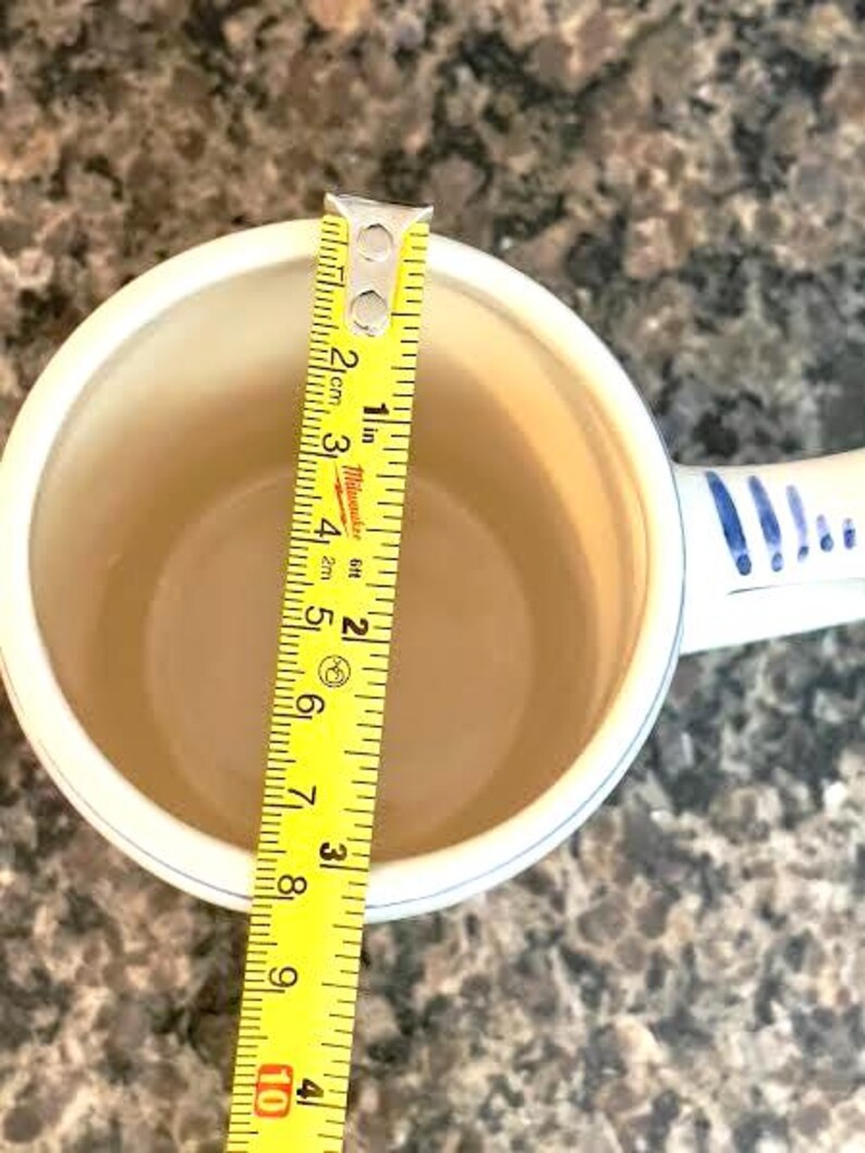 May include: A white ceramic mug with a blue patterned handle and rim, measured with a yellow tape measure. The tape measure shows the mug is approximately 3 inches in diameter.