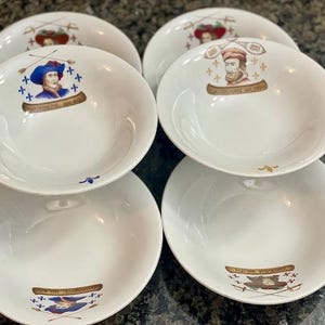 May include: Six white ceramic bowls with decorative illustrations. Each bowl features a different portrait and emblem in the center, with gold and blue accents. The bowls are stacked in pairs, showcasing the detailed designs.