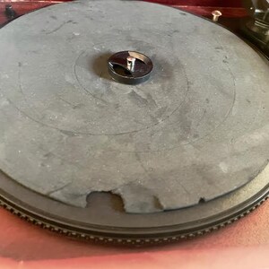 May include: Close-up of a vintage turntable with a dark grey platter and a black centre spindle. The platter has a worn appearance with a section missing. The turntable is set on a wooden surface.