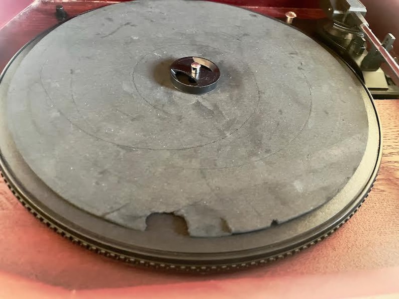 May include: Close-up of a vintage turntable with a dark grey platter and a black centre spindle. The platter has a worn appearance with a section missing. The turntable is set on a wooden surface.
