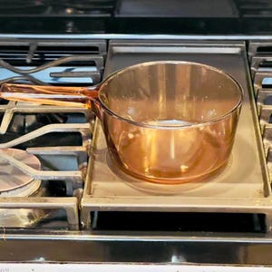 May include: A transparent amber-colored glass saucepan with a single integrated handle. The round cooking vessel features smooth, curved walls and a clear base, designed for stovetop use, showcasing a simple, functional, and minimalist aesthetic.