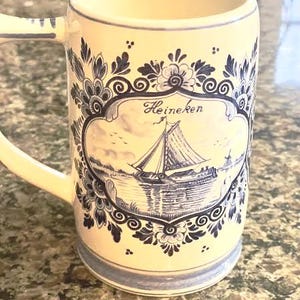 May include: Cream-colored ceramic mug with a handle, featuring a blue and white nautical design. The mug has a decorative border and a central image of a sailboat with the word "Heineken" written above it.