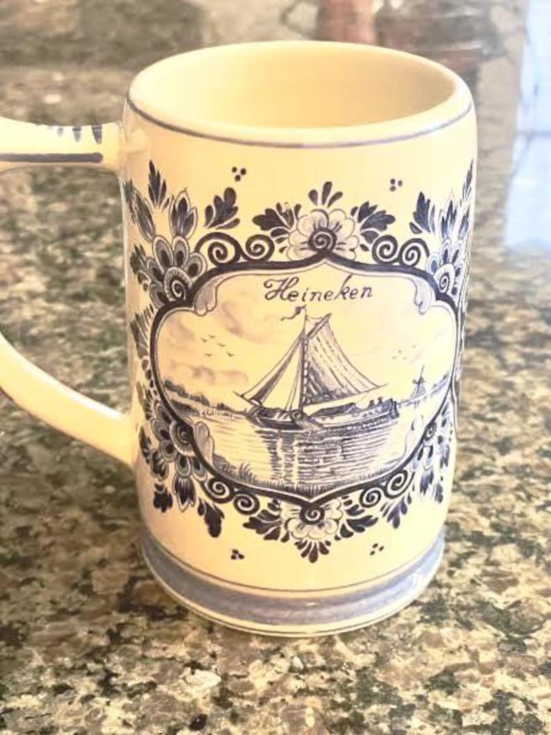 May include: Cream-colored ceramic mug with a handle, featuring a blue and white nautical design. The mug has a decorative border and a central image of a sailboat with the word "Heineken" written above it.