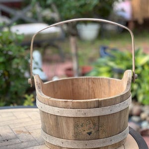 May include: A round wooden bucket with a light brown finish, featuring two horizontal bands and a metal swing handle. The exterior surface displays a small, dark, stenciled graphic of a bird surrounded by floral patterns on the front side.