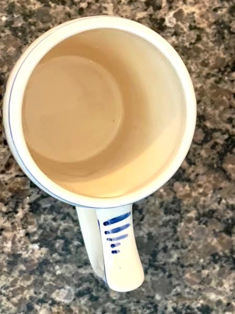 May include: White ceramic mug with a beige interior and a white handle. The handle is decorated with blue vertical lines and dots. The rim of the mug has a thin blue line.