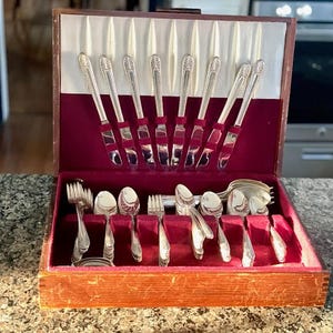 Puede incluir: Un cofre de cubiertos de madera vintage con interior de terciopelo rojo. El cofre está abierto, mostrando un juego completo de cubiertos plateados, que incluyen tenedores, cucharas y cuchillos. Los cuchillos están dispuestos en una fila vertical.