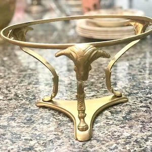 Puede incluir: Un soporte decorativo de metal dorado con una parte superior redonda y un diseño de cabeza de carnero en el centro. El soporte tiene tres patas curvas y una base decorativa, adecuado para exhibir artículos.