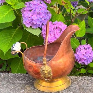 Könnte beinhalten: Ein Kohleeimer aus Kupfer mit einem Messingsockel und einem schwenkbaren Henkel. Der Griff verfügt über ein weißes Keramikstück mit blauen Blumenmustern. Ein Messingornament in Form eines Löwenkopfes ist an der Seite des Gefäßes angebracht.