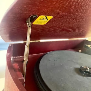 May include: A vintage record player with a dark red wooden casing. The lid is open, showing a black turntable and a yellow sticker with text. A metal hinge is visible, indicating a classic audio device.