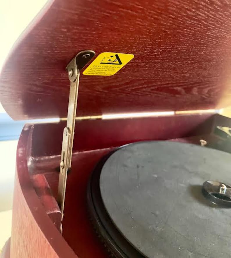 May include: A vintage record player with a dark red wooden casing. The lid is open, showing a black turntable and a yellow sticker with text. A metal hinge is visible, indicating a classic audio device.