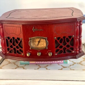 May include: A vintage-style, red wooden radio with a decorative front panel, speaker grilles, and a central dial. The radio features brass-coloured knobs and a clock-style display. The top is a darker wood. The radio is sitting on a white table.