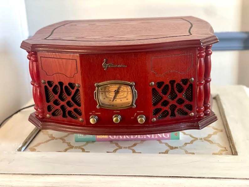 May include: A vintage-style, red wooden radio with a decorative front panel, speaker grilles, and a central dial. The radio features brass-coloured knobs and a clock-style display. The top is a darker wood. The radio is sitting on a white table.