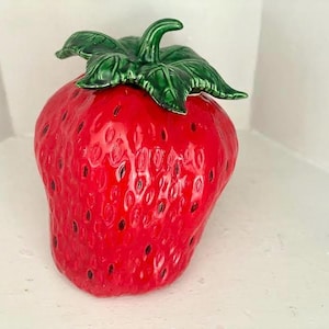 May include: A ceramic strawberry figurine with a glossy red finish and green leaves. The strawberry has a textured surface with small black seeds. The figurine is a decorative item.