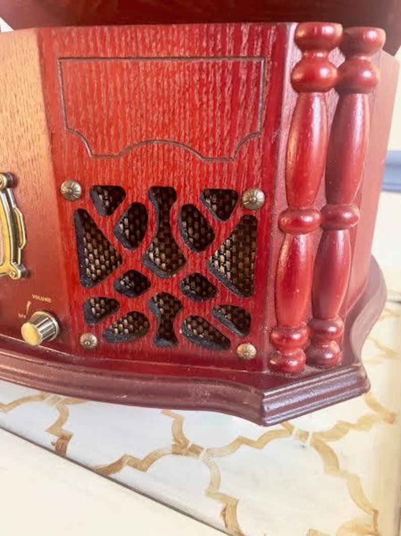 May include: Close-up of a vintage wooden radio with a rich, reddish-brown finish. The radio features decorative wooden accents, including carved details and a series of patterned speaker covers. A volume knob and decorative hardware are visible.