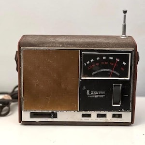 May include: A vintage Zenith Solid State portable radio featuring a brown leather-like casing, a gold-toned speaker grille, and a black control panel. The device includes a silver telescopic antenna, a tuning dial, and a volume slider.