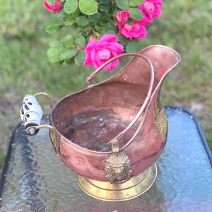 May include: A copper and brass coal scuttle with a decorative lion's head emblem. The handle has a white and blue porcelain grip. The scuttle sits on a brass base. Pink roses are in the background.