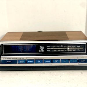 Puede incluir: Un reloj despertador electrónico GE vintage con una parte superior de veta de madera y una pantalla digital. El reloj tiene una esfera plateada y negra con indicadores de frecuencia de radio y botones de control. El texto "ELECTRONIC TOUCH CONTROL" es visible.