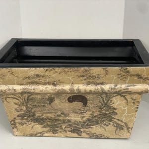 Könnte beinhalten: Rechteckiger Pflanzkübel mit schwarzem Rand und Innenraum. Die Außenseite hat einen cremefarbenen Hintergrund mit einem schwarz-grauen Landschaftsdesign. Der Pflanzkübel hat ein dekoratives, gealtertes Aussehen, geeignet für den Innen- oder Außenbereich.