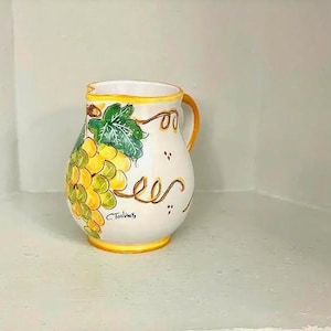 May include: White ceramic jug with a yellow handle and rim, decorated with a cluster of yellow grapes and green leaves. The jug has a curved design and is likely used for serving beverages.