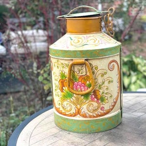 May include: A decorative metal container with a hinged lid and handle. The container is painted with a cream background and features floral and fruit designs in orange, pink, and green. The top and bottom edges are painted green.