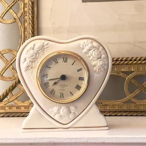 May include: A heart-shaped, off-white ceramic clock with a gold-tone bezel and Roman numeral clock face. The clock is decorated with rose motifs and sits on a white surface. Decorative gold frames are in the background.