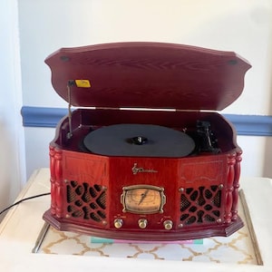 Puede incluir: Un tocadiscos de madera roja de estilo vintage con tapa abatible. El tocadiscos tiene un plato negro, un dial dorado y rejillas de altavoz. Se encuentra sobre una mesa blanca con una superficie estampada.