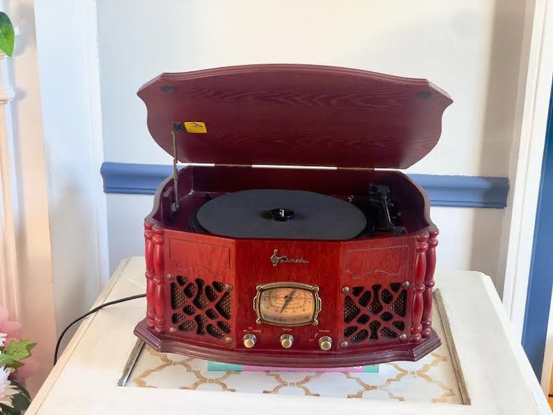 May include: A vintage-style, red wooden record player with a hinged lid. The record player has a black turntable, a gold-toned dial, and speaker grilles. It sits on a white table with a patterned surface.